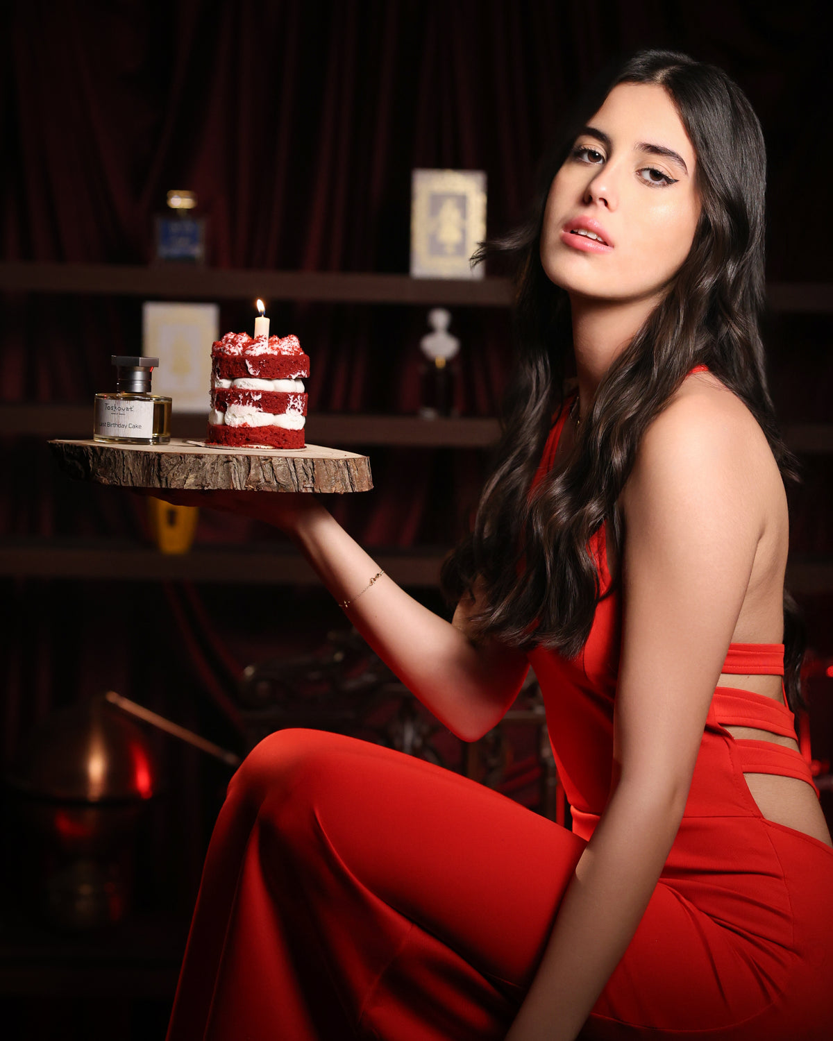 Toskovat Last Birthday Cake bottle and a candlelit red velvet cake on a wood slice, held up by a woman in a red jumpsuit, gourmand niche perfume decant
